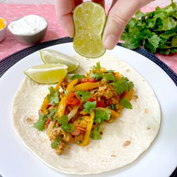 cilantro lime chicken fajitas on a flour tortilla with a hand squeezing lime juice over it. Lime wedges, cilantro, sour cream and cheese on the side.
