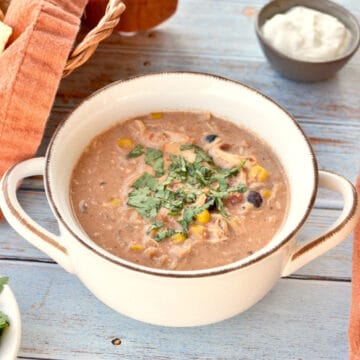 chicken chili made in crockpot with cream cheese, beans, corn, tomatoes, chilis, spices, and cilantro. Chili is inside a white bowl, with chips, sour cream and cilantro on the side.