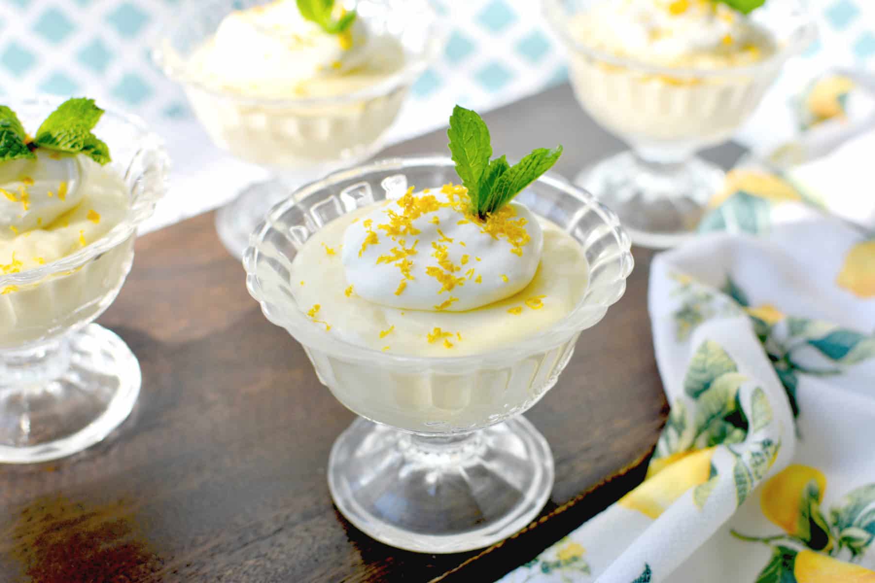 homemade lemon pudding in cups topped with whipped cream, lemon zest and mint leaves.