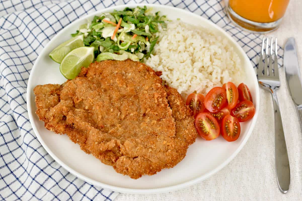 Milanesa steak on a plate served with grape tomatoes, white rice, salad, and lime edges.