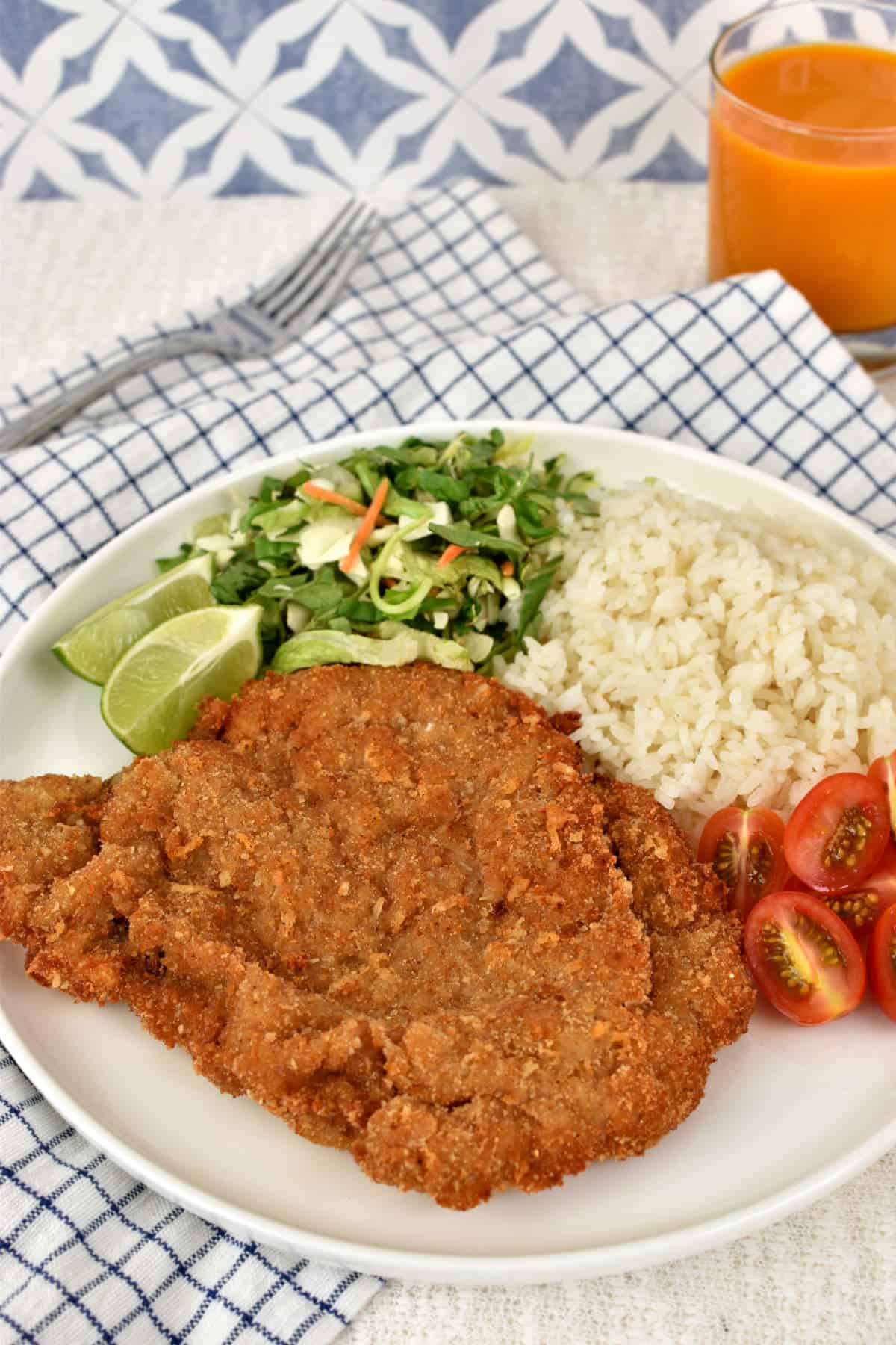 Beef Milanese on a plate served with rice, tomatoes, salad, and lime wedges.
