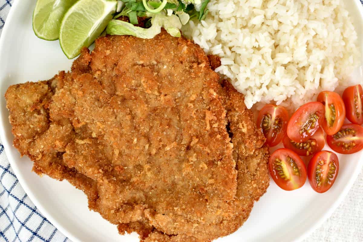 Beef milanesa on a plate with white rice, grape tomatoes, salad, and lime wedges.