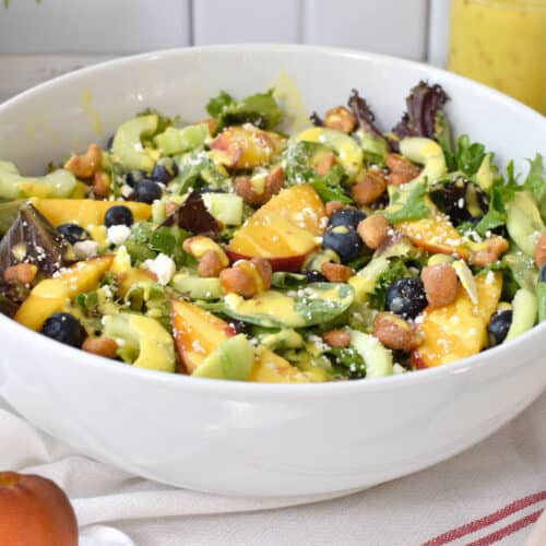 Salad bowl with spring mix salad, peach, feta, blueberries, cucumber, and nuts, topped with mango vinaigrette.