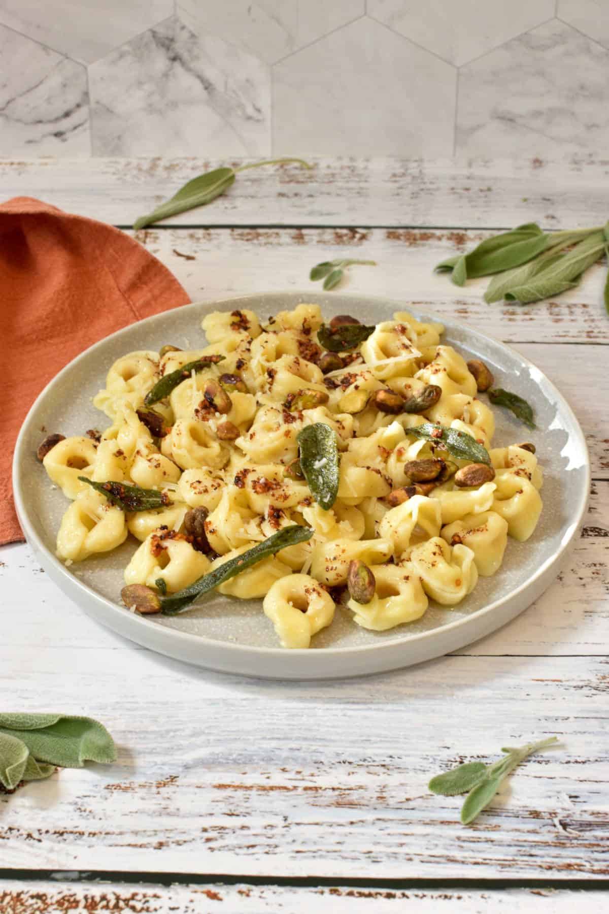A plate with tortellini tossed with butter sauce and topped with parmesan cheese, pistachios and crispy sage leaves.